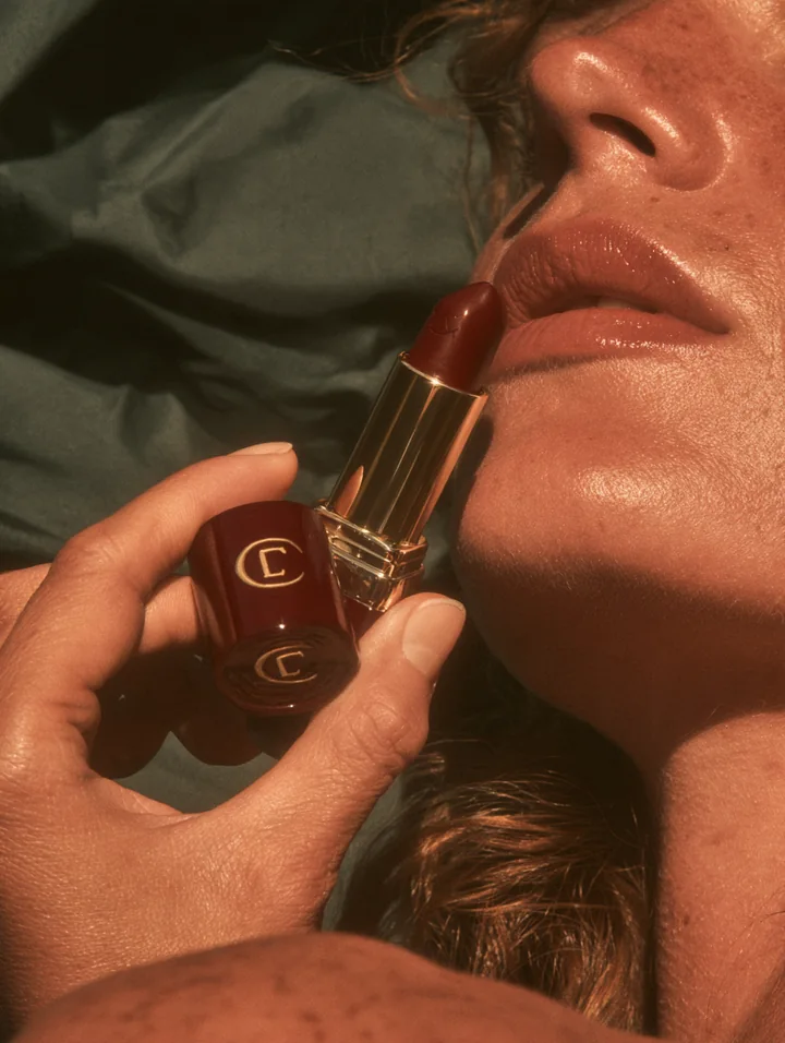 Close-up of a woman holding a deep red lipstick beside her lips in golden sunlight
