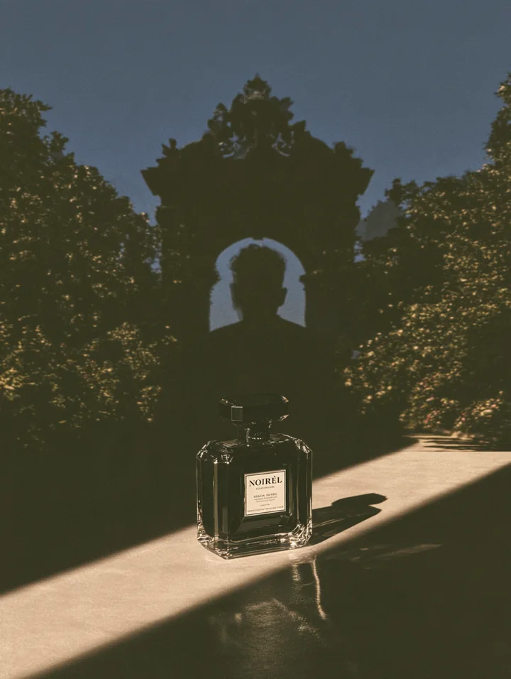 A glass perfume bottle in a shaft of light beneath a monumental silhouette and garden arch
