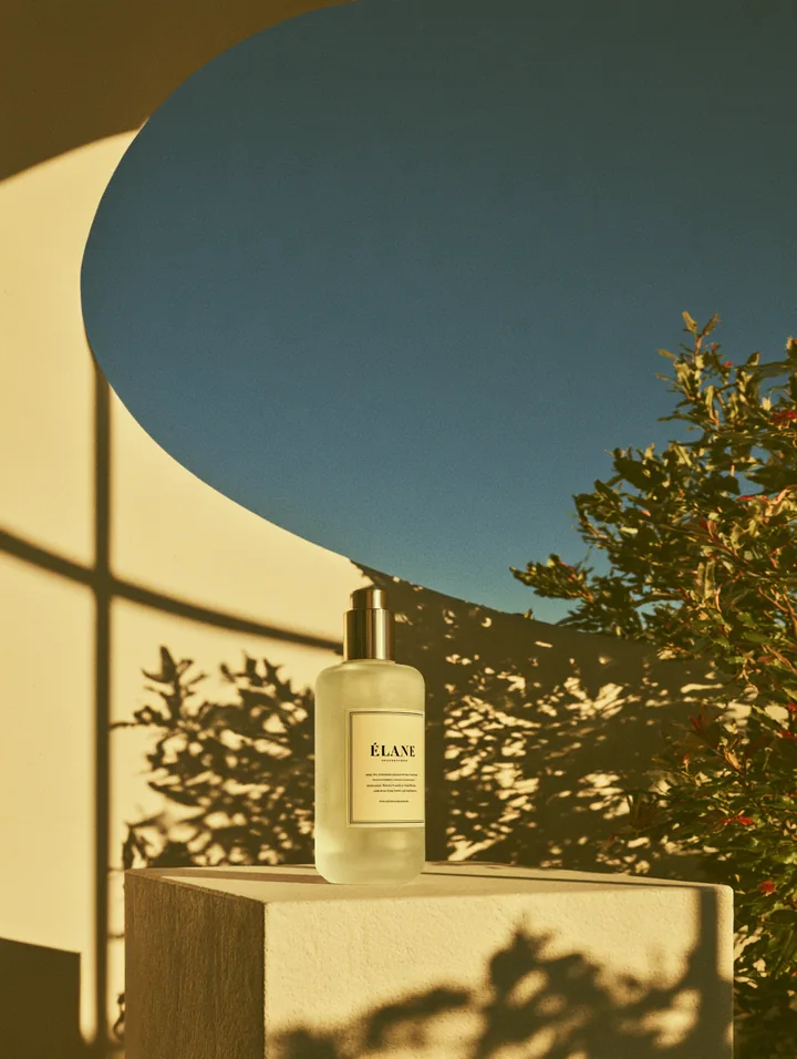 A frosted skincare pump bottle on a pale plinth beneath curved architecture and blue sky